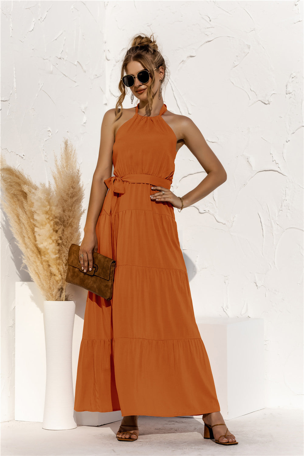 Model is wearing halter neck tiered maxi dress with tie front waist in the color terracotta.